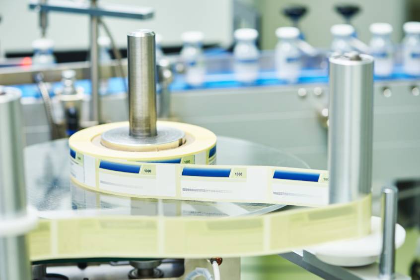 Automated labeling machine applying product labels to vials in a pharmaceutical packaging line.