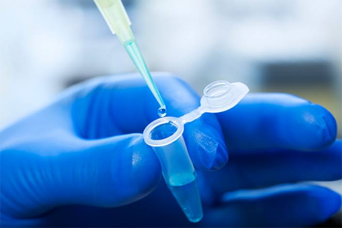 Gloved hand holding a microcentrifuge tube while a pipette dispenses a liquid sample into it in a laboratory setting.