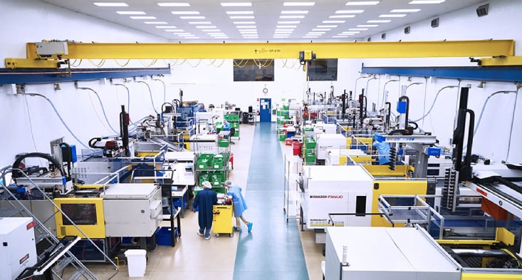 Brightly lit injection molding production floor with multiple machines, overhead crane system, and technicians in cleanroom attire operating medical manufacturing equipment.