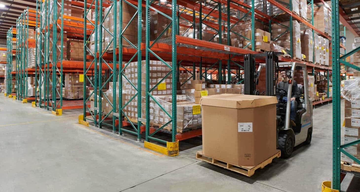 Warehouse with tall storage racks filled with labeled boxes, as a forklift operator transports a large packed carton on a wooden pallet.